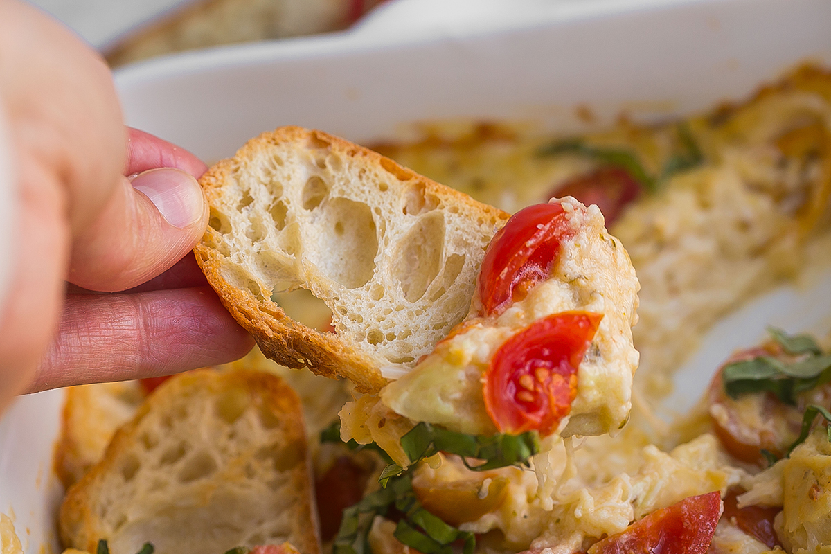 hand holding slice of bread dipped into hot caprese dip